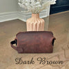 Dark brown leather toiletry bag on a wooden floor with a vase of flowers in the background.