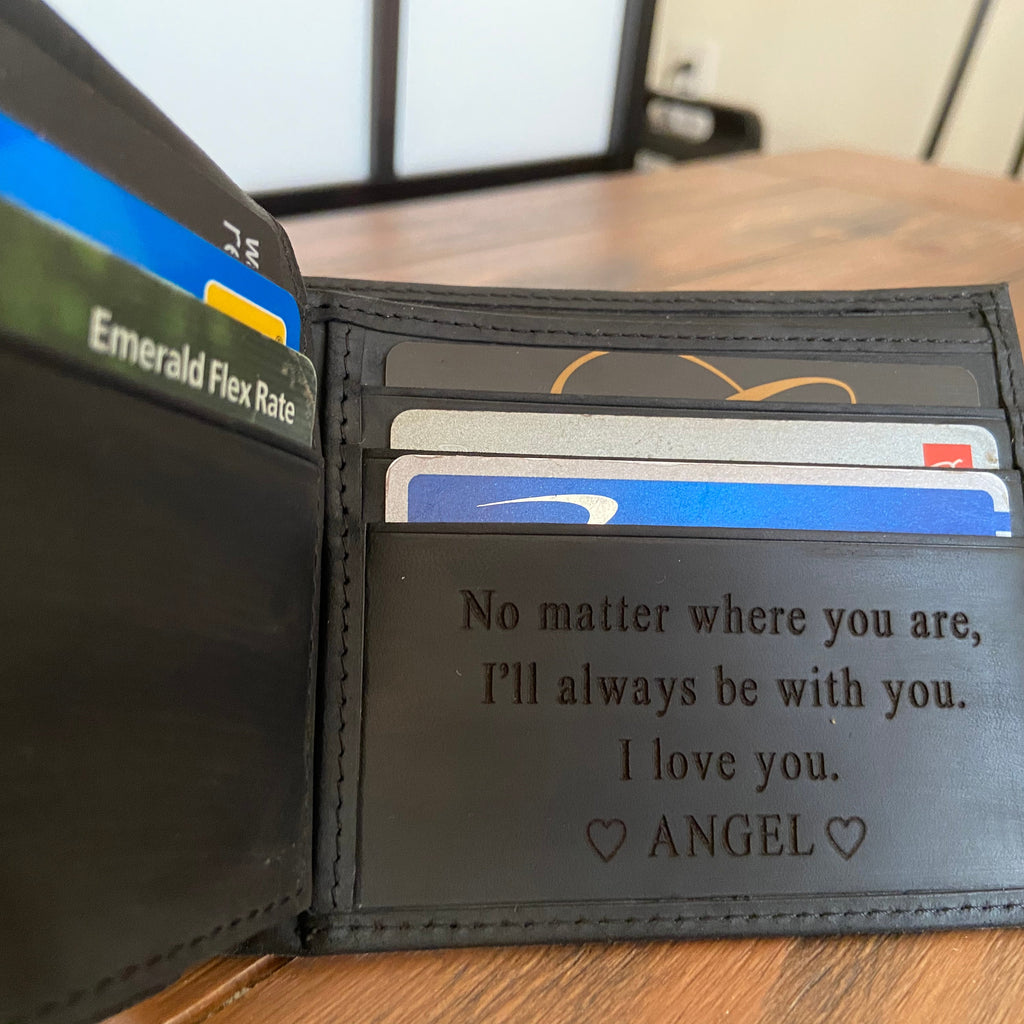 A black leather bifold wallet shown open on a wooden surface, with card slots holding several cards and a printed message inside: 'No matter where you are, I'll always be with you. I love you.