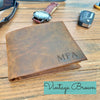 Brown leather wallet with 'MFA' initials on a wooden surface, surrounded by sunglasses, a watch, and car keys.