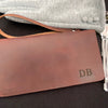 A close-up of a brown leather checkbook or wallet with an embossed 'DB' monogram, resting on a black textured surface with a gray fabric item partially visible behind it.
