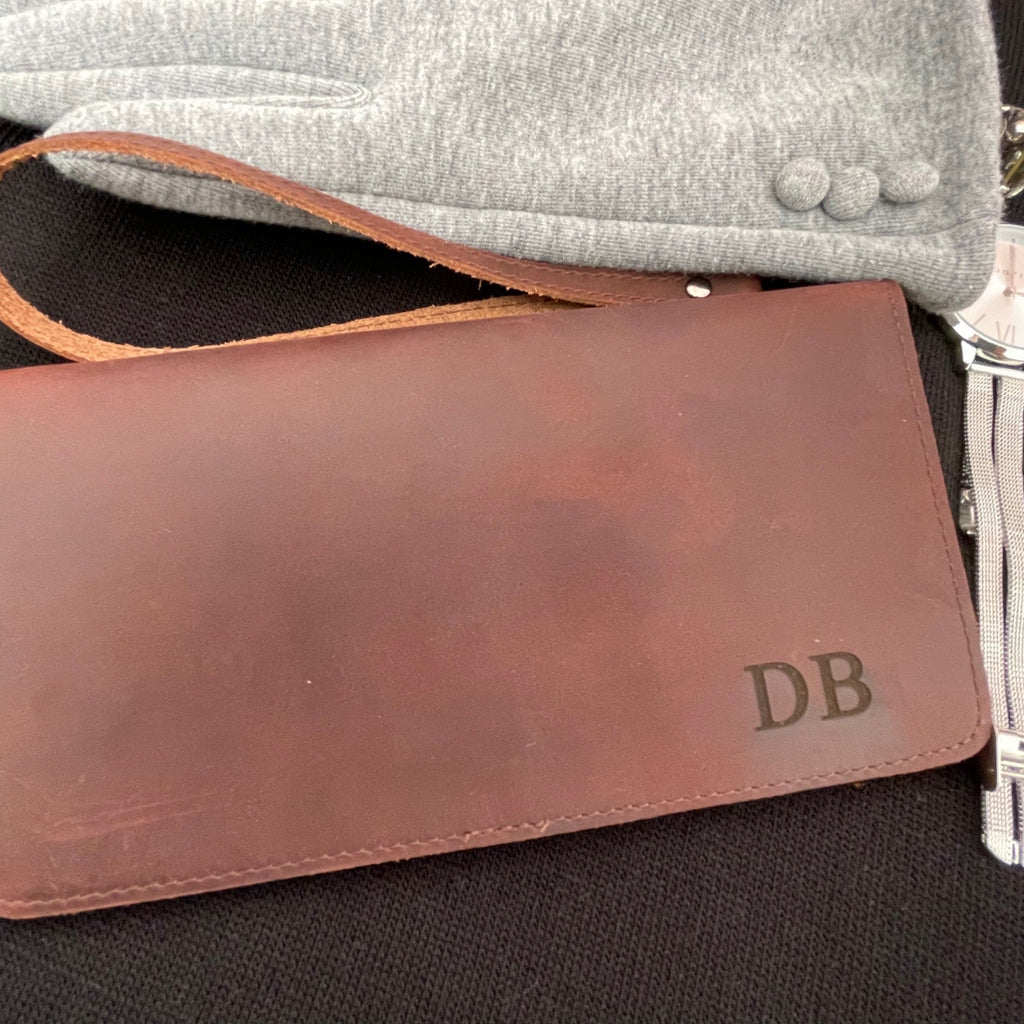 A close-up of a brown leather checkbook or wallet with an embossed 'DB' monogram, resting on a black textured surface with a gray fabric item partially visible behind it.