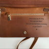 A close-up of a brown leather wallet or clutch showing the interior side with a zipper pocket, metal snap button, and printed sentimental text that reads, 'Work days can be long but years are short. We will win the lottery someday.