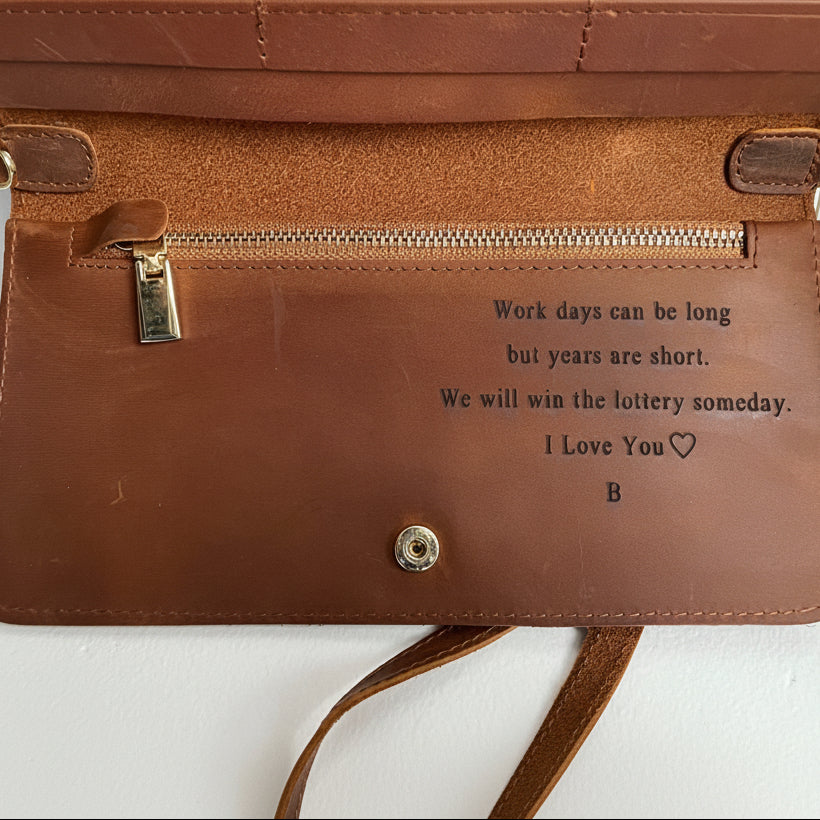 A close-up of a brown leather wallet or clutch showing the interior side with a zipper pocket, metal snap button, and printed sentimental text that reads, 'Work days can be long but years are short. We will win the lottery someday.