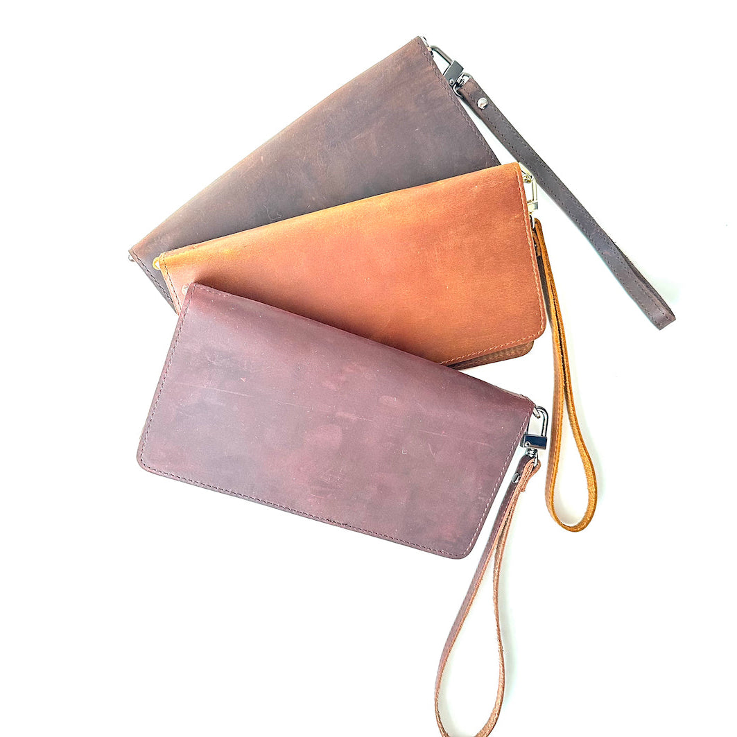 Three leather women's wallets with wrist straps, laid out flat against a white background. They come in brown, black, and tan colors.